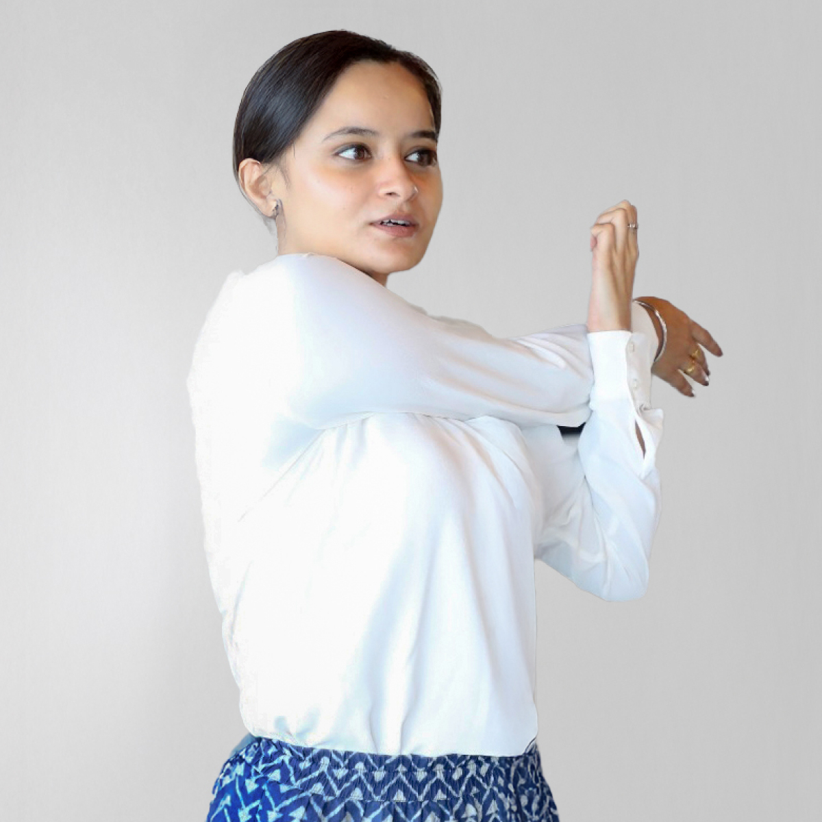 Woman stretching her arm wearing a white shirt and blue patterned skirt, standing against a plain light background